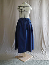 Load image into Gallery viewer, The back view of an ankle-length dark royal blue skirt in a crisp woven fabric, with pleats at the narrow waistband, on a dressform in a studio. There's a bow made from the waist ties at the centre back.