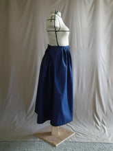 Load image into Gallery viewer, The right side view of an ankle-length dark royal blue skirt in a crisp woven fabric, with pleats at the narrow waistband, on a dressform in a studio. There's a deep pleat in the side where the front of the skirt overlaps the back, and we see a bow made from waist ties peeking out at the back waist.
