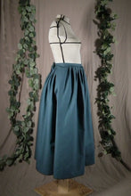 Load image into Gallery viewer, The side view of an ankle-length forest green skirt in a crisp woven fabric, with pleats at the narrow waistband, on a dressform in a studio. There's a deep pleat in the side where the front of the skirt overlaps the back, and we see a bow made from waist ties peeking out at the back waist. There are also some fake green vines in the background.