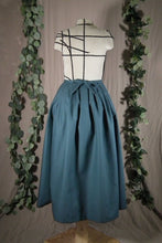 Load image into Gallery viewer, The back view of an ankle-length forest green skirt in a crisp woven fabric, with pleats at the narrow waistband, on a dressform in a studio, with some fake green vines in the background. There's a bow made from the waist ties at the centre back.