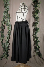 Load image into Gallery viewer, The side view of an ankle-length black skirt in a drapey fabric, with gathers at the waistband, on a dressform in a studio. The ties from the waist make a bow at the centre back. There are some fake green vines in the background.