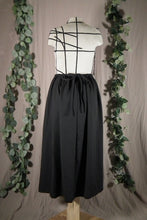Load image into Gallery viewer, The back view of an ankle-length black skirt in a drapey fabric, with gathers at the waistband, on a dressform in a studio. The ties from the waist make a bow at the centre back. There are some fake green vines in the background.