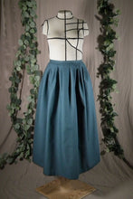 Load image into Gallery viewer, The front view of an ankle-length forest green skirt in a crisp woven fabric, with pleats at the narrow waistband, on a dressform in a studio, with some fake green vines in the background.