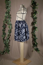Load image into Gallery viewer, A short knee-length elastic waistband skirt in a drapey rayon print that's dark blue with white flowers is displayed on a dress form, showing the side view. In the background are some fake green vines.