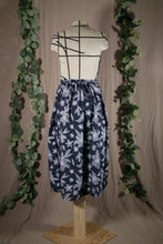 Load image into Gallery viewer, The back view of An ankle-length pleated skirt with waits ties, in a drapey rayon print that's dark blue with white flowers is displayed on a dress form. There’s a bow made from the ties at the centre back. In the background are some fake green vines.