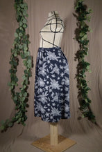 Load image into Gallery viewer, The side view of an ankle-length pleated skirt with waits ties, in a drapey rayon print that's dark blue with white flowers is displayed on a dress form. In the background are some fake green vines.