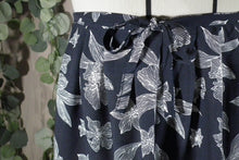 Load image into Gallery viewer, A close-up view of the pleated waistband of a skirt in a drapey rayon print that's dark blue with white flowers, displayed on a dress form, showing a cute bow at the waist. There are some blurry fake vines in the background