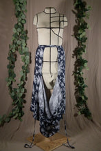 Load image into Gallery viewer, The open front view of an ankle-length pleated skirt with waits ties, in a drapey rayon print that's dark blue with white flowers, on a dressform in a studio, with some fake green vines in the background. The front part of the skirt has fallen open to reveal the waist ties from the back portion of the skirt.