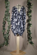 Load image into Gallery viewer, A long pleated skirt in a drapey rayon print that's dark blue with white flowers is displayed on a dress form as a swim coverup, showing the back view. The slit makes a deep V at the neck, and ties make drapey bows at the shoulder. In the background are some fake green vines.