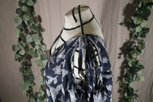 Load image into Gallery viewer, A birds eye view of a drapey rayon print skirt that's dark blue with white flowers, displayed on a dress form as a swim coverup, showing off the ties making bows on the shoulder. The slit makes a deep V at the neck. In the background are some fake green vines.