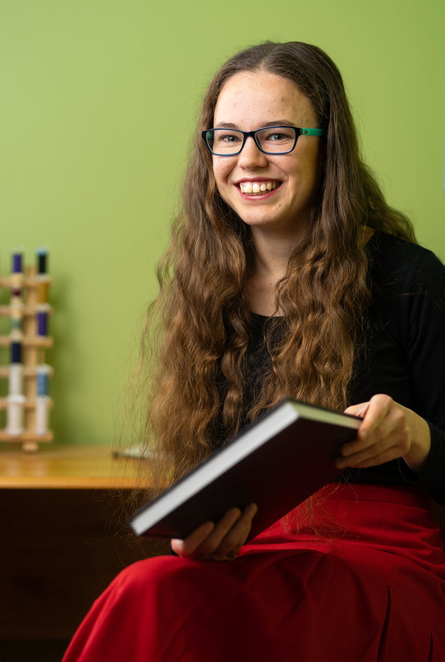 A young woman with long wavy brown hair smiles at the camera. She wears glasses and holds a sketchbook. The spring green background contrasts with her bright red skirt. In the background, a thread spool holder sits on a desk. 