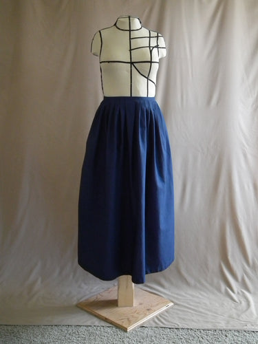 The front view of an ankle-length dark royal blue skirt in a crisp woven fabric, with pleats at the narrow waistband, on a dressform in a studio.