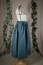 Load image into Gallery viewer, The side view of an ankle-length forest green skirt in a crisp woven fabric, with pleats at the narrow waistband, on a dressform in a studio. There&#39;s a deep pleat in the side where the front of the skirt overlaps the back, and we see a bow made from waist ties peeking out at the back waist. There are also some fake green vines in the background. 
