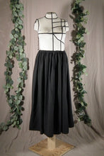 Load image into Gallery viewer, The front view of an ankle-length black skirt in a drapey fabric, with gathers at the waistband, on a dressform in a studio. There are some fake green vines in the background.
