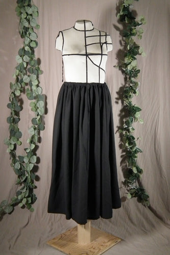 The front view of an ankle-length black skirt in a drapey fabric, with gathers at the waistband, on a dressform in a studio. There are some fake green vines in the background.