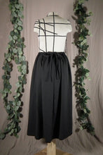 Load image into Gallery viewer, The back view of an ankle-length black skirt in a drapey fabric, with gathers at the waistband, on a dressform in a studio. The ties from the waist make a bow at the centre back. There are some fake green vines in the background.
