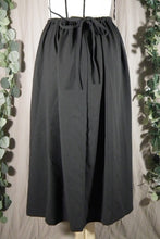 Load image into Gallery viewer, A closer back view of an ankle-length black skirt in a drapey fabric, with gathers at the waistband, on a dressform in a studio. The picture is over-exposed to show the details of the gathers and bow. The ties from the waist make a bow at the centre back. There are some fake green vines in the background.
