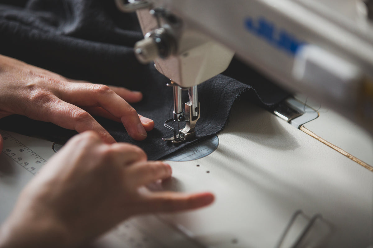 Hands run dark grey fabric through an industrial sewing machine. 