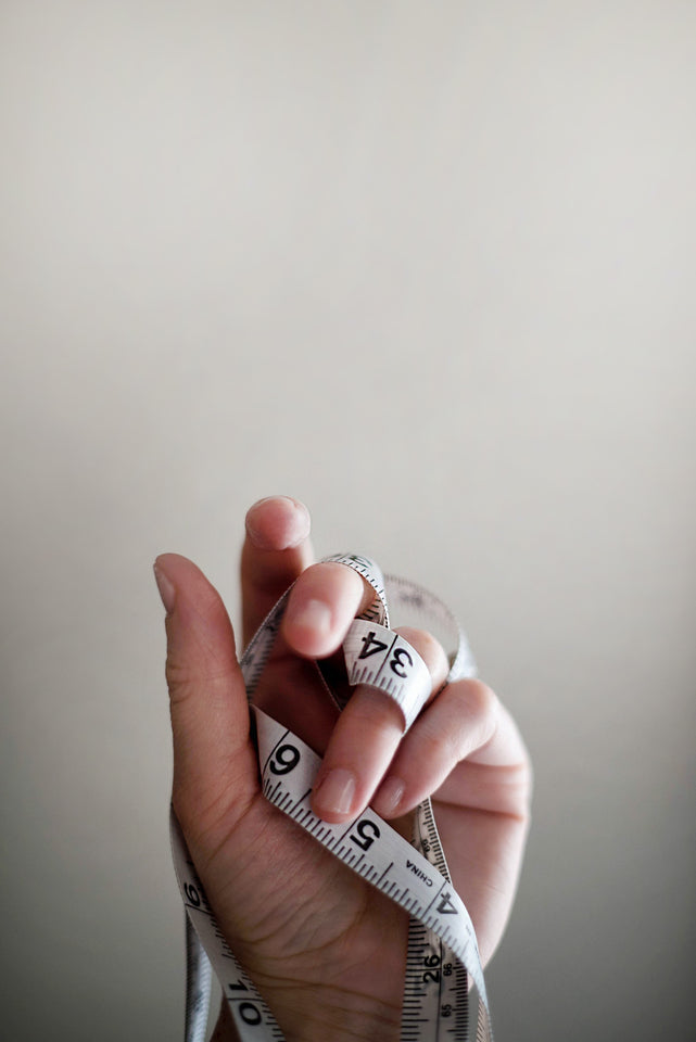 A hand entwined with a measuring tape fills the lower half of the photo, against a pale backdrop. 