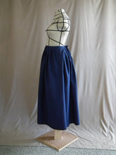 Load image into Gallery viewer, The left side view of an ankle-length dark royal blue skirt in a crisp woven fabric, with pleats at the narrow waistband, on a dressform in a studio. There&#39;s a deep pleat in the side where the front of the skirt overlaps the back, and we see a bow made from waist ties peeking out at the back waist. 
