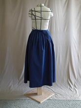 Load image into Gallery viewer, The back view of an ankle-length dark royal blue skirt in a crisp woven fabric, with pleats at the narrow waistband, on a dressform in a studio. There&#39;s a bow made from the waist ties at the centre back.

