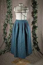 Load image into Gallery viewer, The front view of an ankle-length forest green skirt in a crisp woven fabric, with pleats at the narrow waistband, on a dressform in a studio, with some fake green vines in the background.

