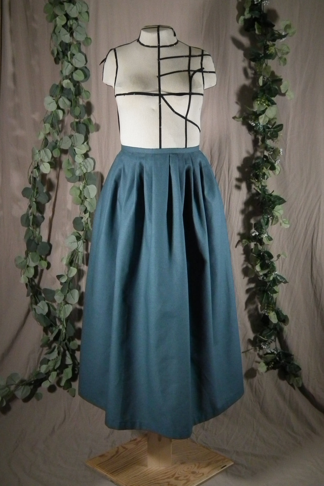 The front view of an ankle-length forest green skirt in a crisp woven fabric, with pleats at the narrow waistband, on a dressform in a studio, with some fake green vines in the background.