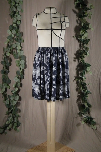 A short knee-length elastic waistband skirt in a drapey rayon print that's dark blue with white flowers is displayed on a dress form, showing the front view. In the background are some fake green vines.