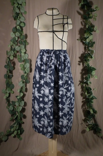 An ankle-length pleated skirt with waits ties, in a drapey rayon print that's dark blue with white flowers is displayed on a dress form, showing the front view. There’s a bow tied at the centre front. In the background are some fake green vines.